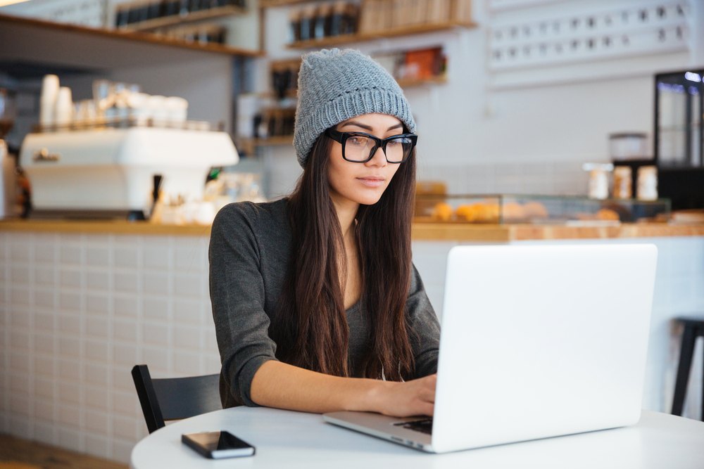 trendy woman wearing beanie and glasses working on laptop in coffee shop